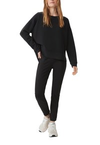 Czarny sweatshirt o luźnym kroju i szerokich rękawach, zestawiony z czarnymi zwężanymi spodniami. Model w białych trampkach. Miękkie tekstury, minimalistyczny design.