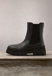 Bottine noire en cuir avec semelle épaisse et panneau latéral élastique sur surface en bois sur fond uni.