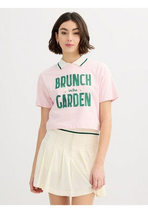 Mujer con pelo corto y castaño que lleva una camiseta polo rosa corta con el texto "Brunch in the Garden" y una falda plisada crema.