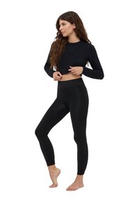 Maglione nero a cropped in morbido materiale, abbinato a leggings neri con una texture liscia e una vita alta. Modella in piedi a piedi nudi.