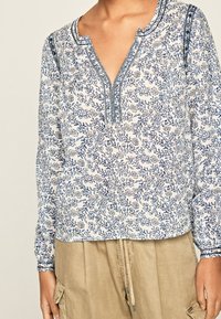 Blusa a fiori bianca e blu, con scollatura a V e maniche lunghe con polsini elasticizzati. Tessuto morbido con cuciture dettagliate.