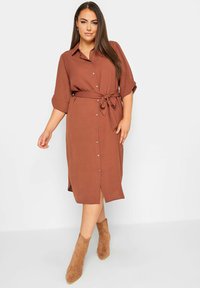 Braunes Hemdkleid mit Taillengurt, kurzen Ärmeln und mittellanger Länge. Der Stoff sieht glatt aus und hat einen leichten Glanz. Wildleder-Stiefeletten. 