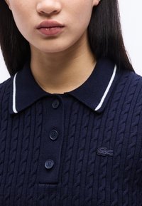 Νεαρό άτομο που φοράει μπλε σκούρο πλεκτό πόλο με τρία κουμπιά και μικρό κεντημένο λογότυπο κροκόδειλου στο στήθος.