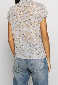 Top floral azul claro con mangas cortas, que presenta un tejido texturizado, pequeños patrones de flores amarillas y marrones, y un diseño de espalda plizada.