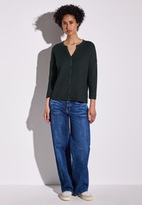 Mörkgrön skjorta med knappar och trekvart ärm, i kombination med blå vida jeans och ljusa sneakers mot en neutral bakgrund.