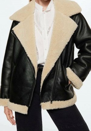 Veste d'aviateur en cuir noir avec col, poignets et ourlet en shearling crème portée sur un chemisier blanc et un pantalon noir.