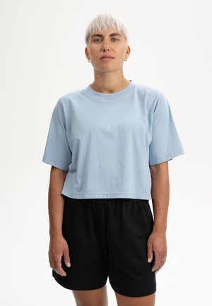 Person mit kurzen platinblonden Haaren, der ein hellblaues, bauchfreies T-Shirt und schwarze Shorts trägt, vor einem weißen Hintergrund steht.