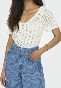 Top de punto blanco con mangas cortas que presenta un patrón texturizado y un escote en V amplio, combinado con unos jeans de talle alto en tono azul jaspeado.
