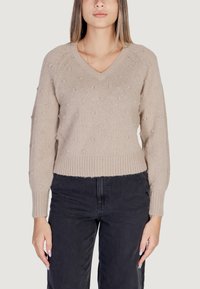 Maglione beige a maglia con scollatura a V, pompon texturizzati e polsini e orlo a coste. Abbinato a pantaloni neri a vita alta.