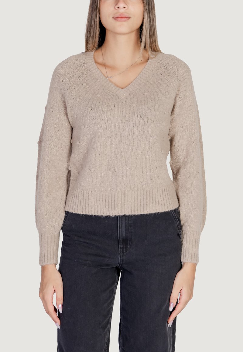 Maglione beige a maglia con scollatura a V, pompon texturizzati e polsini e orlo a coste. Abbinato a pantaloni neri a vita alta.