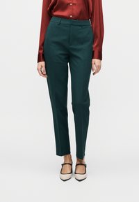 Pantalon vert sur mesure à coupe droite, avec une texture lisse et un pli avant marqué, associé à une chemise en satin rouge.