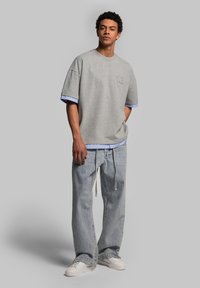 Gray short-sleeve sweatshirt with a blue and white striped hem, paired with light blue baggy jeans and white sneakers. Accent logo on the chest.