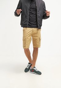 Czarna kurtka bomberka na wzorzystym czarnym t-shircie, khaki szorty cargo i zielone trampki z czerwonymi i białymi akcentami.