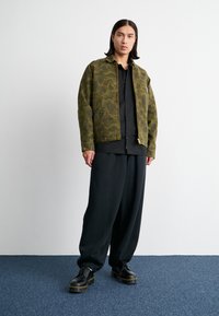 Veste bomber verte olive avec un motif camouflage, portée sur une chemise noire à boutons et un pantalon noir ample, complétée par des chaussures noires.