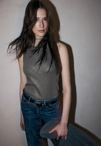 Bez rękawów, metalicznie szary top w połączeniu z niebieskimi dżinsami i czarnym paskiem. Naturalne włosy opadają luźno, a w jednej ręce trzyma szary element garderoby.