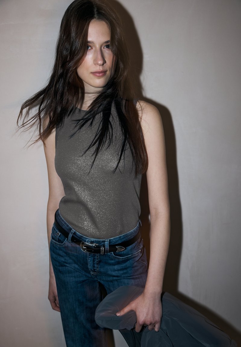 Bez rękawów, metalicznie szary top w połączeniu z niebieskimi dżinsami i czarnym paskiem. Naturalne włosy opadają luźno, a w jednej ręce trzyma szary element garderoby.