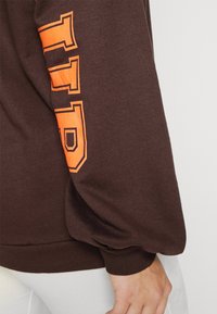 Manica di felpa marrone con lettere "TTO" contornate di arancione, indossata da una persona con pelle chiara e pantaloni bianchi, su uno sfondo chiaro.