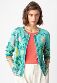 Un cárdigan de punto turquesa con patrones florales en rosa, amarillo y verde, con acentos decorativos en los puños y un cuello redondo.