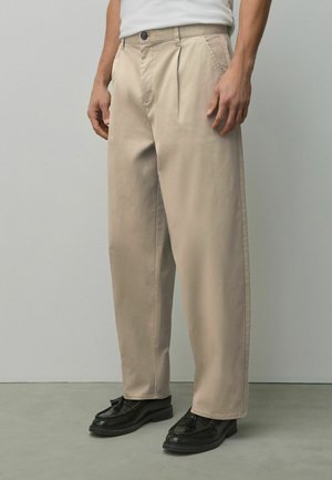 Man wearing beige pleated trousers and black tassel loafers, standing on gray floor against a plain light gray wall.