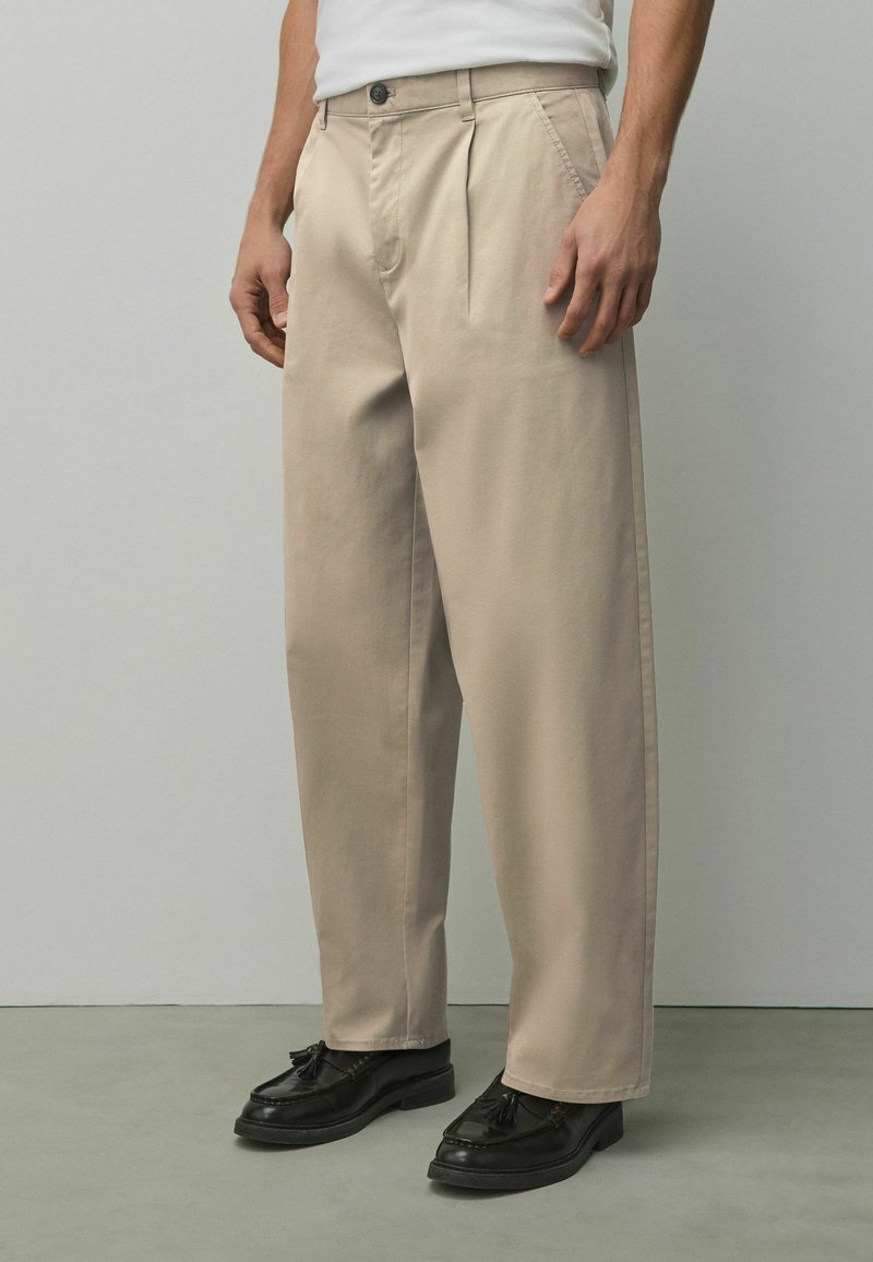 Homme portant un pantalon beige plissé et des mocassins noirs à pampilles, debout sur un sol gris devant un mur uni gris clair.