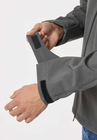 Personne ajustant la manchette d'une veste grise avec une fermeture à scratch, gros plan sur les mains et la manche sur un fond uni.