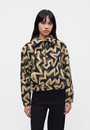 Jeune femme aux cheveux lisses et foncés porte un pull duveteux avec des motifs zigzag jaunes et violets et un pantalon noir, debout devant un fond uni.