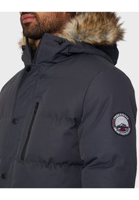 Grå pufferjakke med hætte foret med pels, med en lynlåslomme på brystet og et rundt logo-patch på venstre ærme. Læg i kviltet tekstur.