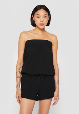 Ung kvinde med kort sort hår iført en sort stropløs romper, stående med afslappede arme mod en ensfarvet grå baggrund.