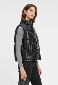Schwarze Leder-Puffweste mit hohem Kragen, Reißverschluss vorne und zwei großen Fronttaschen. Gefütterter Stoff mit glatter Textur.