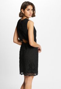 Zwarte kanten jurk met mouwloos ontwerp, bloemenpatroon, uitgesneden rug en gegolfde zoom. De gestructureerde stof voegt diepte toe aan de algehele uitstraling.