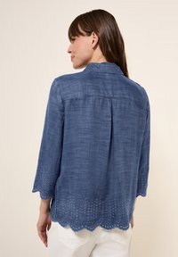 Blå linneskjorta med trekvartslånga ärmar, scallopad nederkant med öljettbroderi och en avslappnad passform. Bakvy utan synliga knappar.