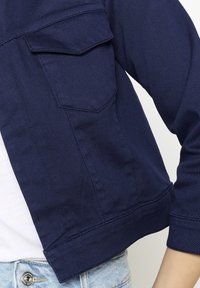 Granatowa krótka kurtka z dwoma kieszeniami na piersi, wykonana z gładkiego materiału, z lekkim połyskiem i podwiniętymi rękawami, idealna na codzienne stylizacje.