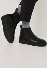 Schwarze Lederstiefeletten mit elastischen Seiteneinsätzen, strukturierten Gummisohlen und minimalistischen Design, getragen mit grauen Socken und schmal zulaufenden Hosen.
