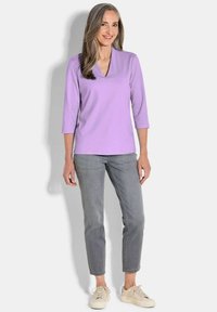 Lichtpaarse V-hals top met driekwartmouwen, gecombineerd met grijze slim-fit jeans en beige sneakers, tegen een witte achtergrond.