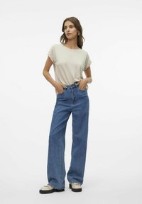 Top beige a maniche corte abbinato a jeans blu a vita alta e gamba larga. Le scarpe sono di un crema chiaro con suole nere. Tessuto liscio, vestibilità casual.