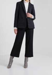 Blazer tailored in misto lana di un grigio scuro con due tasche frontali, indossato sopra una blouse bianca testurizzata, abbinato a pantaloni neri a gamba larga e stivaletti grigi alla caviglia.