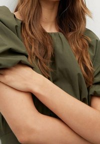 Olivgrünes Stoffoberteil mit Puffärmeln und weitem Ausschnitt. Haare offen, Arme verschränkt, mit glattem Hautton und -struktur.