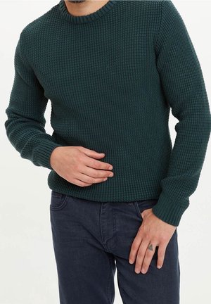 Mand iført mørkegrøn tekstureret sweater og mørke jeans, med den ene hånd hvilende på maven og den anden ved siden, der viser en lille trekantet tatovering.