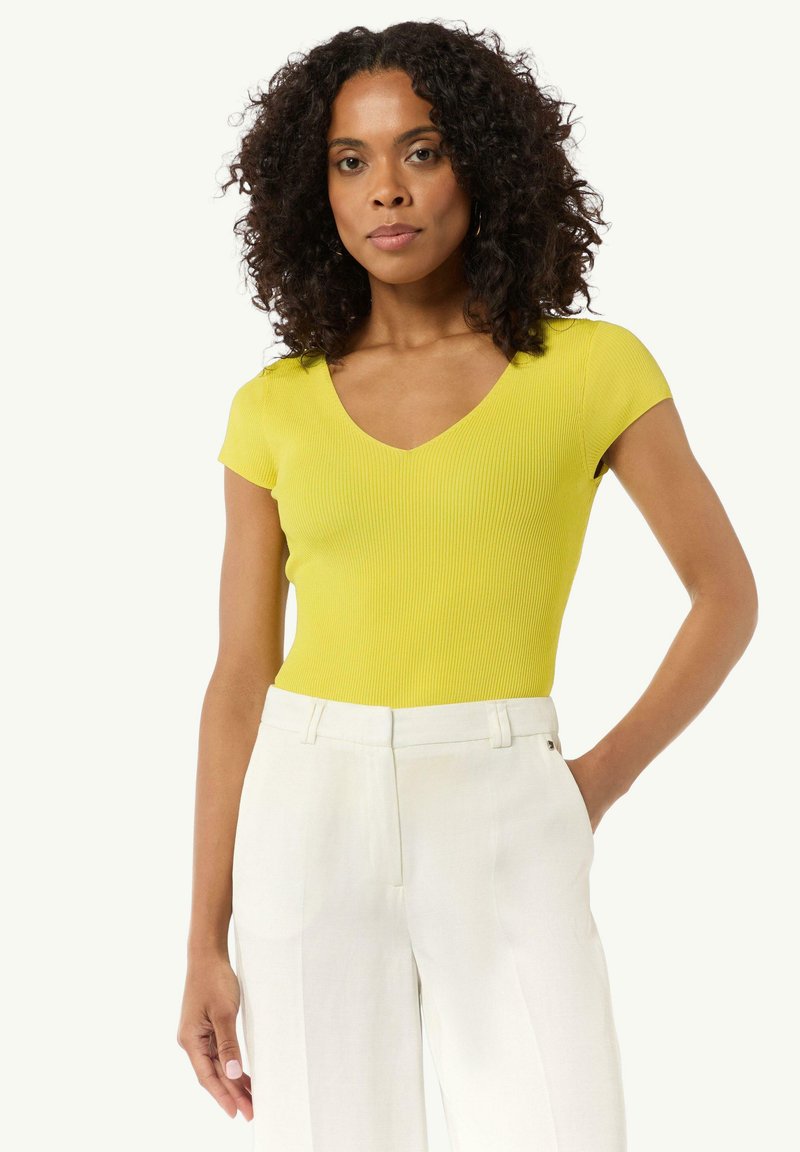 Żółty, prążkowany bodysuit z krótkim rękawem i dekoltem w kształcie V, w zestawieniu z białymi, lnianymi spodniami o wysokim stanie. Widoczne detale struktury i dopasowania.