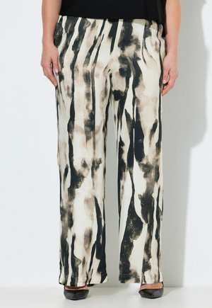 Frau trägt lockere, hoch taillierte Hosen mit vertikalen schwarzen und beigen abstrakten Streifen, kombiniert mit schwarzen spitzen Schuhen.