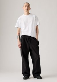 Mand med tatoveringer iført en simpel hvid t-shirt, sorte cargo-bukser og sorte sko, stående foran en neutral baggrund.