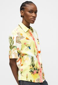 Νεαρό άτομο με πλεξούδες που φοράει κοντομάνικο πουκάμισο με κουμπιά διακοσμημένο με απεικονίσεις πουλιών, κάκτων, καπέλων και κραγιόν.