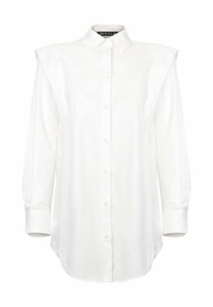 Balti marškiniai su traškiu apykakle, ilgomis rankovėmis ir petnešėmis. Lygus audinys su šiek tiek struktūruota forma ir apvaliu kraštu.