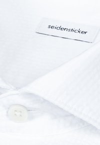 Col de chemise texturée blanche avec un bouton blanc et une étiquette de marque "seidensticker" à l'intérieur.