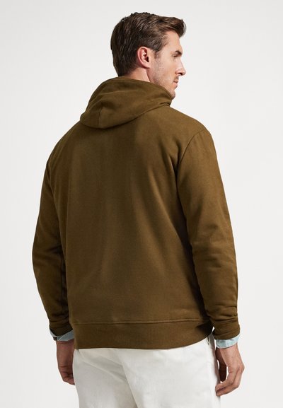 Άνδρας που φοράει καφέ φούτερ με κουκούλα πάνω από ανοιχτό μπλε πουκάμισο και λευκό παντελόνι, γυρισμένος πλάτη σε απλό λευκό φόντο.