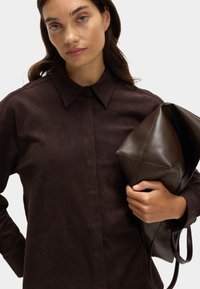 Hnědá semišová košile s límcem na knoflíky, spárovaná s tmavě hnědou koženou kabelkou s geometrickým tvarem a měkkou texturou.