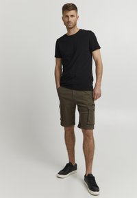 Czarny t-shirt z krótkim rękawem w zestawieniu z oliwkowymi szortami cargo z kieszeniami, noszony z czarnymi trampkami na neutralnym tle.