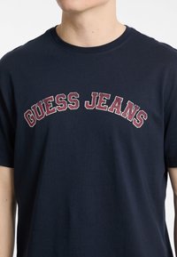 Tamna plava pamučna majica s natpisom "GUESS JEANS" u kontrastnim burgundskim i bijelim slovima preko prsnog dijela. Kratki rukavi.