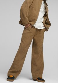 Hnědá fleecová bunda a kalhoty s volným střihem. Pod tím bílý tričko. Černé a oranžové tenisky se šněrováním. Měkká textura po celém povrchu.