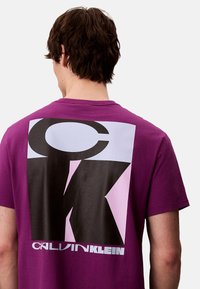 T-shirt en coton violet arborant un grand motif graphique au dos avec les lettres "CK" en noir et rose clair, accompagné du texte "CALVIN KLEIN".